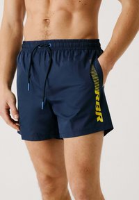 Bañador navy con cintura elástica, cordón y un gráfico lateral en amarillo. El material es suave y resistente al agua.