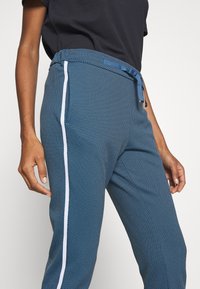 Osoba ubrana w niebieskie spodnie w wzory z białym paskiem po boku i niebieskim sznurkiem, w zestawieniu z czarną koszulą z krótkim rękawem.