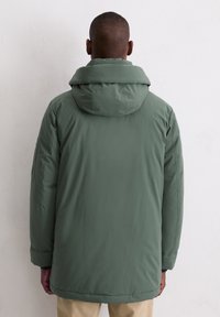 Man die met zijn rug naar de camera staat, gekleed in een groene gewatteerde winterjas en beige broek, tegen een witte muur.