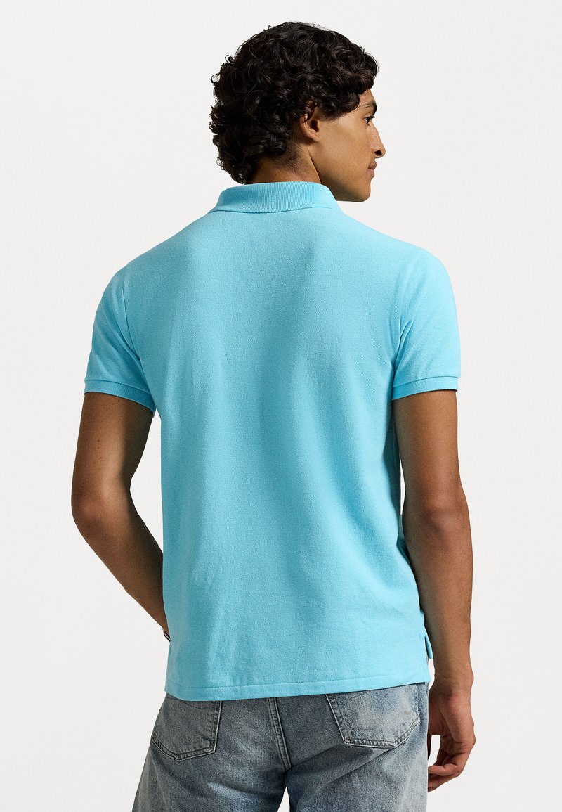 Mann mit lockigem Haar, der ein hellblaues kurzärmeliges Polo-Shirt und ausgewaschene blaue Jeans trägt, mit dem Rücken zu einer schlichten Hintergrundkulisse.