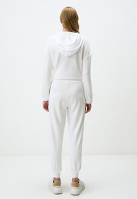 Witte, zachte katoenen trainingspak met een capuchon, voorzien van manchetten aan de mouwen en de enkels voor een snug fit. Draagt met witte sneakers.
