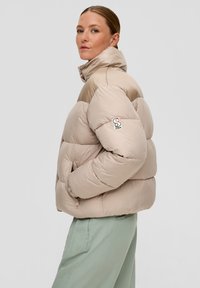 Beige pufīgs mētelis ar augstu apkakli, quilted dizainu un izšūtu logo uz piedurknes. Iespējas ir rāvējslēdzēja kabatas un mājīga tekstūra.