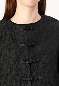 Black textured jacket featuring a wavy fabric, round neckline, and three knot-style closures on the front. Long sleeves are not visible.