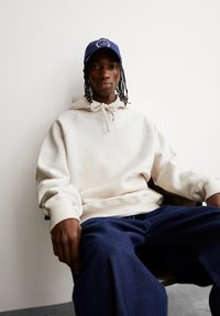 Felpa beige chiaro con cappuccio e coulisse, con un piccolo logo, abbinata a pantaloni larghi blu scuro, indossata con un cappello blu navy.