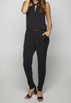 Femme portant une combinaison noire sans manches avec un col licou, des poches latérales, une ceinture fine, et des sandales noires à talons blocs ouvertes à l'avant.