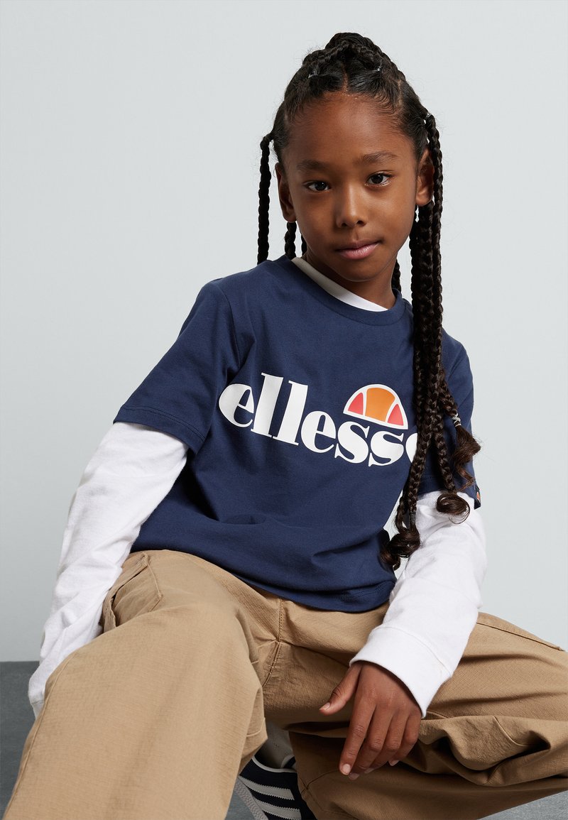 Dunkelblauses T-Shirt mit weißem Logo, über einem weißen Langarmshirt getragen. Beigefarbene, weite Hose. Lange geflochtene Haare. Einfacher Hintergrund.