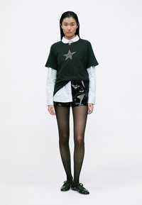 Tricou negru cu un grafic de stea gri, suprapus peste o cămașă albă. Şorturi negre lucioase, ciorapi negri transparenți și pantofi slip-on negri. Fundal simplu.