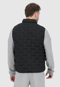 Černá péřová vesta s prošívaným vzorem, vysokým límcem a hladkým povrchem, nošená přes šedou mikinu, z pohledu ze zad.