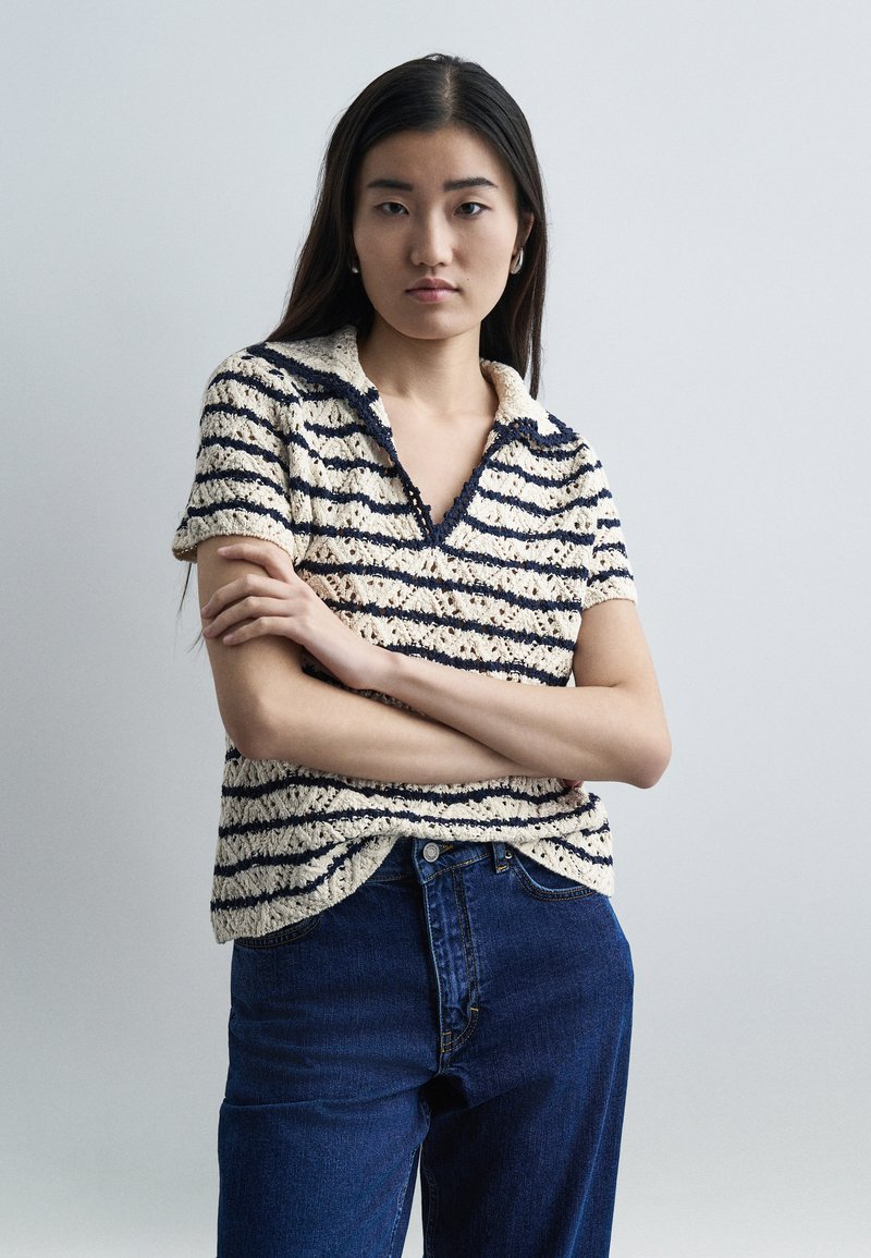 Γυναίκα με μακριά σκούρα μαλλιά, φοράει μπεζ και ναυτικό ριγέ κοντομάνικη πλεκτή μπλούζα και μπλε τζιν, στέκεται με σταυρωμένα τα χέρια μπροστά σε απλό φόντο.