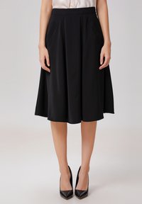 Jupe noire mi-longue en A, fabriquée dans un tissu lisse, avec des poches latérales et une taille ajustée. Associée à des talons noirs pointus.