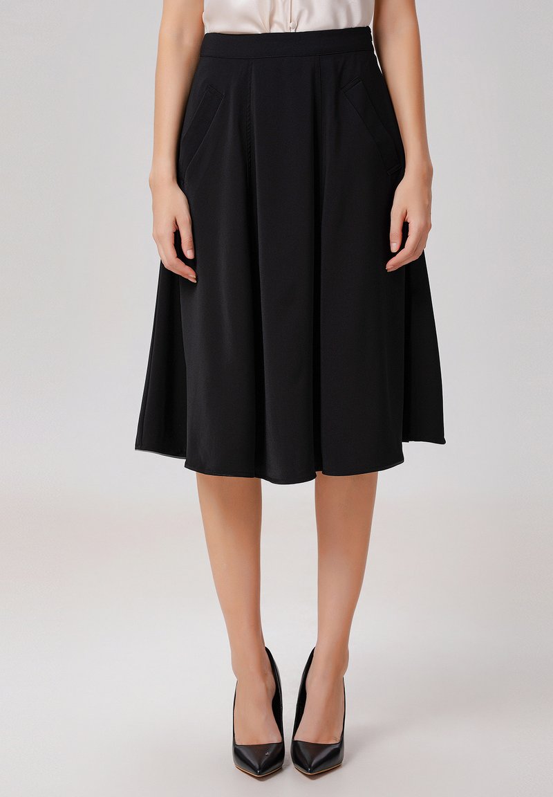 Jupe noire mi-longue en A, fabriquée dans un tissu lisse, avec des poches latérales et une taille ajustée. Associée à des talons noirs pointus.