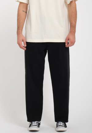 Pantalons noirs, amples, en denim avec une texture lisse. Portés avec un t-shirt clair et ample et des baskets noires.