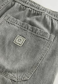 Pantaloni scurți din denim gri cu un tiv elastic, prevăzuți cu un buzunar pe spate și o etichetă de marcă pătrată cu un design solar. Țesătură texturată cu cusături.