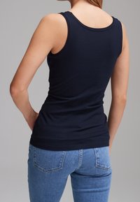 Femme portant un débardeur bleu marine ajusté et un jean denim bleu clair, debout les mains sur les hanches, dos à la caméra.