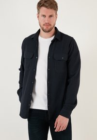 Camicia a righe blu navy con chiusura a bottoni, due tasche sul petto e maniche lunghe. Indossata sopra una maglietta bianca a girocollo, tessuto testurizzato.