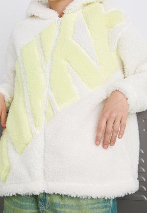 Personne portant une veste sherpa blanche zippée avec de grandes lettres jaune pâle et un jean vert, la main posée sur la hanche.