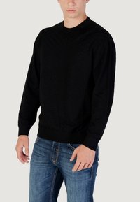 Maglione crewneck nero con texture, maniche lunghe, polsini e orlo a coste, indossato con jeans blu. Motivo sottile sulla superficie del tessuto.