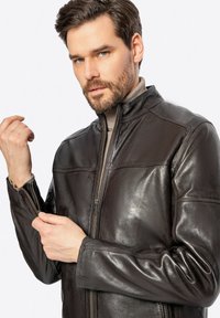 Giubbotto in pelle marrone con zip frontale, colletto alto e dettagli di cucitura visibili. Il materiale ha una consistenza liscia e lucida.