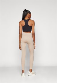 Top corto negro con tirantes tipo racerback, leggings de tiro alto beige acanalados y zapatillas blancas, vistos desde la parte posterior contra un fondo claro y liso.
