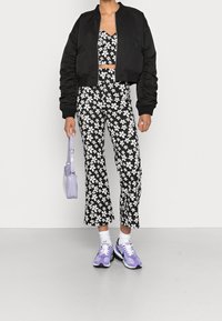 Persona con chaqueta corta negra, blusa de flores en blanco y negro y pantalones, sosteniendo un bolso lavanda y usando zapatillas lavanda y blancas.