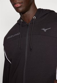 Crna zip-up dukserica s kapuljačom, sivi Mizuno logo na prsima, izrađena od mekog materijala s bijelim detaljima duž bokova.