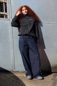 Sort letvægtsjakke med lynlåse, parret med løse blå denimjeans. Baggrunden har et metalpanel med et gittervindue.