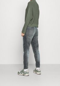Olivgrön stickad tröja, grå denimjeans med en urtvättad finish, och gröna sneakers med vita accenter. Bakifrån visas den avslappnade passformen och den avslappnade stilen.
