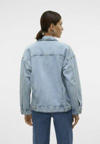 Svetlo modra denim jakna sproščenega kroja z vidnim šivanjem in gumbnimi detajli, ujemajoča se z modrimi kavbojkami. Gladka tekstura s klasičnim dizajnom.