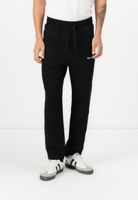 Black sweatpants with elastic waistband, drawstring, and side pockets; lightweight fabric; embroidered logo on the left thigh; tapered fit.