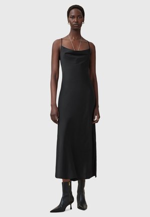 Abito lungo in satin nero con spalline sottili, scollatura a cowl discreta e spacco laterale; abbinato a stivali neri alla caviglia con cerniera d'oro.