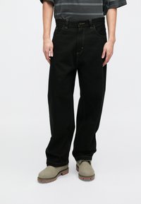 Jeans de mezclilla negra con corte recto, diseño tradicional de cinco bolsillos, detalles de costura amarilla, llevados con botas de ante gris.