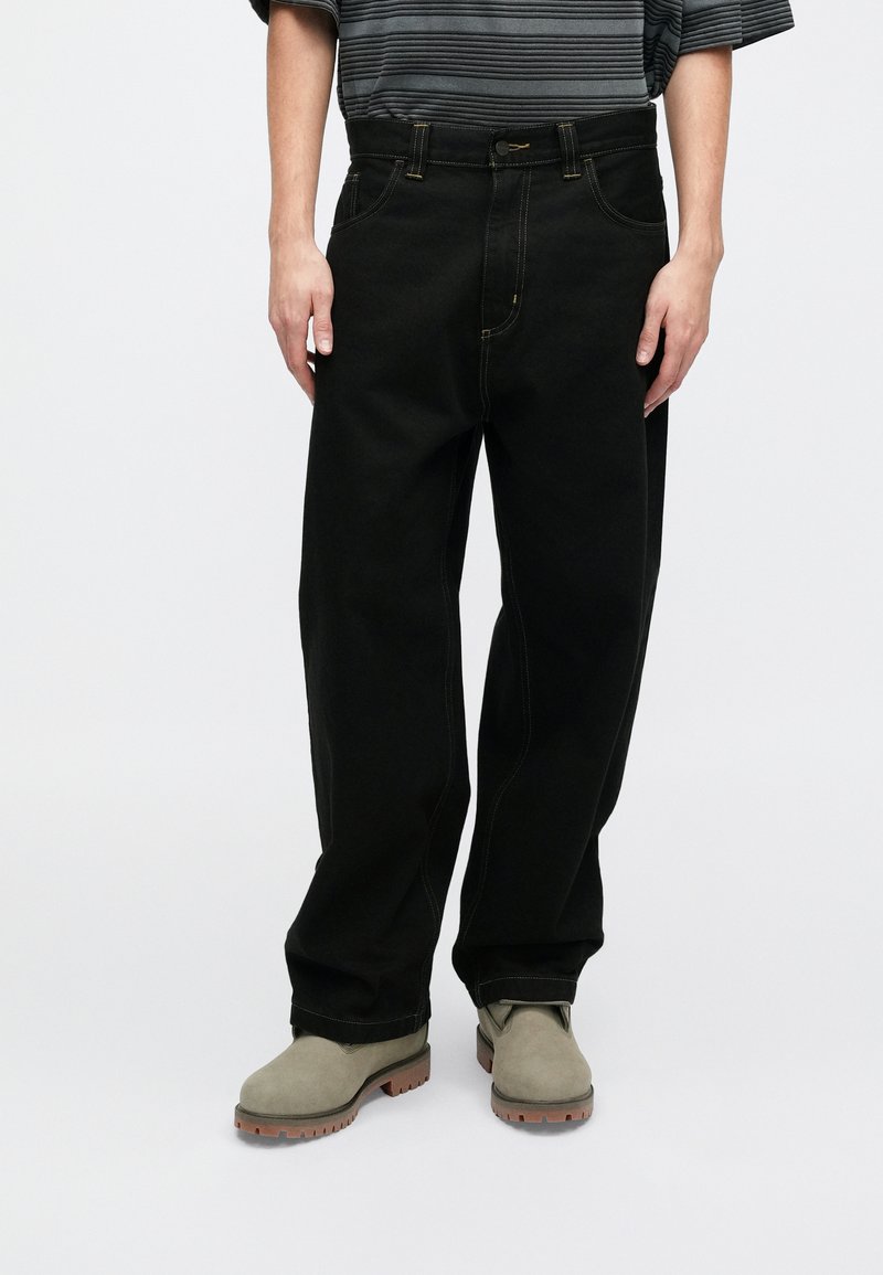 Schwarze Denim-Jeans mit geradem Schnitt, traditionellem Fünf-Taschen-Design, Akzenten in gelben Nähten, kombiniert mit grauen Wildlederstiefeln.