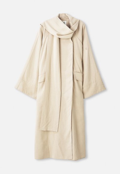 Bežog kut od lagane tkanine s prozračnim dizajnom i širokim rukavima. Ima veliki okovratnik i dva prednja džepa.