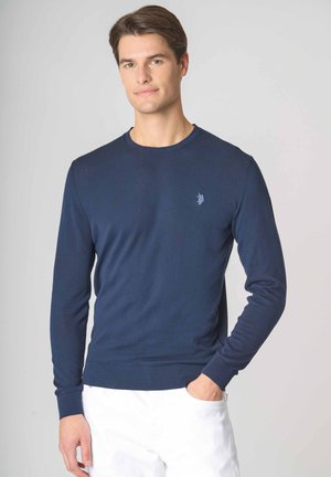 Giovane uomo che indossa un maglione a maniche lunghe blu navy e pantaloni bianchi, in piedi con una mano in tasca contro uno sfondo semplice.