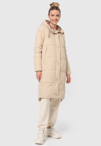 Beige gevoerde lange jas met een capuchon, voorzien van een ritszak en drukknoopsluitingen. Zachte textuur en rechte snit, gecombineerd met lichte sweatpants en sneakers.