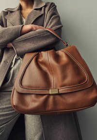 Sac en cuir marron avec une forme décontractée, doté d'un accent en métal doré et d'une texture douce, accompagné d'un manteau et d'un pull gris.