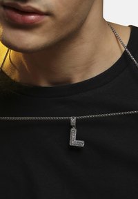 Sølv kæde med vedhæng i stiliseret "L", prydet med klare rhinsten, båret mod en sort skjorte.