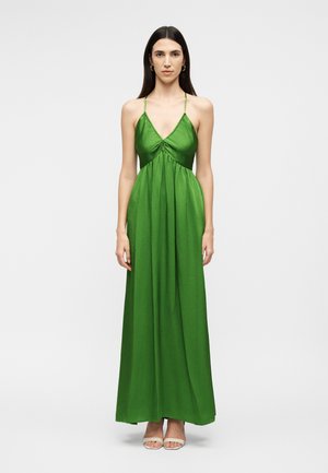 Moteris su ilgais tamsiais plaukais, dėvinti ilga žalią suknelę be rankovių su plonais petnešėliais ir baltus atvirus sandalėlius, stovinti baltame fone.