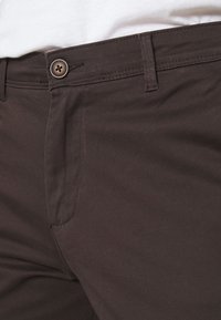 Ruskeat kangashousut, joissa on sileä pinta, yksi metallinen nappikiinnitys ja klassinen vyötärölinja. Huomaa etutaskun yksityiskohta.