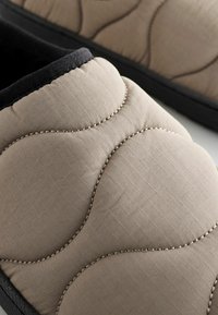 Pantuflas de tela beige con un diseño acolchado en patrón de cuadrícula, borde negro y suela de goma texturizada. Los detalles de costura añaden un toque especial.