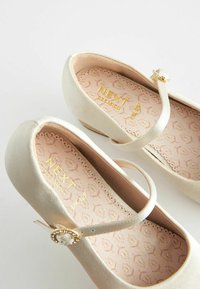 Ballerines couleur crème avec une texture lisse, une bride ornée d'une boucle dorée décorative et des semelles intérieures à motif floral.