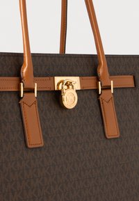 Bolso de tela marrón con estampado, asas de cuero, herrajes dorados y un detalle de cierre con el logo de la marca. Textura suave y forma estructurada.