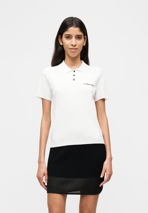 Femme aux longs cheveux foncés portant un polo blanc à manches courtes avec des boutons noirs et une jupe côtelée noire, devant un fond uni.