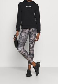 Sudadera negra con capucha y un logo, combinada con leggings grises de estampado animal. También son visibles unas zapatillas deportivas negras y una botella de agua.