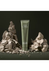 Tube vert étiqueté "Clay Multi-Cleanser" par Doctor Babor, posé sur des fragments de pierre texturée de couleur claire, sur un fond vert foncé.