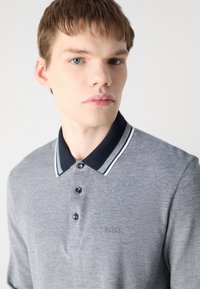 Camisa polo masculina em tecido cinza, com colarinho preto com riscazinhas brancas, placket com três botões e pequeno logo bordado no peito.