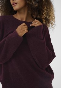 Burgundijos spalvos per didelis megztinis, pagamintas iš minkštos, tekstūruotos megztinės medžiagos su raukšlėtomis rankovėmis. Siluetas turi plačią iškirptę ir laisvą prigludimą.