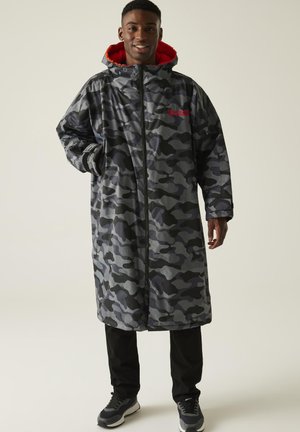 Man glimlachend, gekleed in een lange grijs-zwarte camouflage jas met een rode capuchon voering, zwarte broek en donkere sneakers, staand tegen een effen achtergrond.