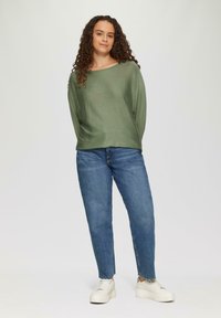 Grüner Strickpullover mit weitem Ausschnitt, kombiniert mit blauen Denim-Jeans. Das Model trägt weiße Plateausneaker. Der Hintergrund ist schlicht und hell.