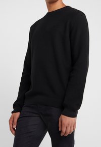 Black, ribbed knit sweater with a crew neckline and long sleeves, featuring subtle logo detailing on the chest. Worn with dark jeans.